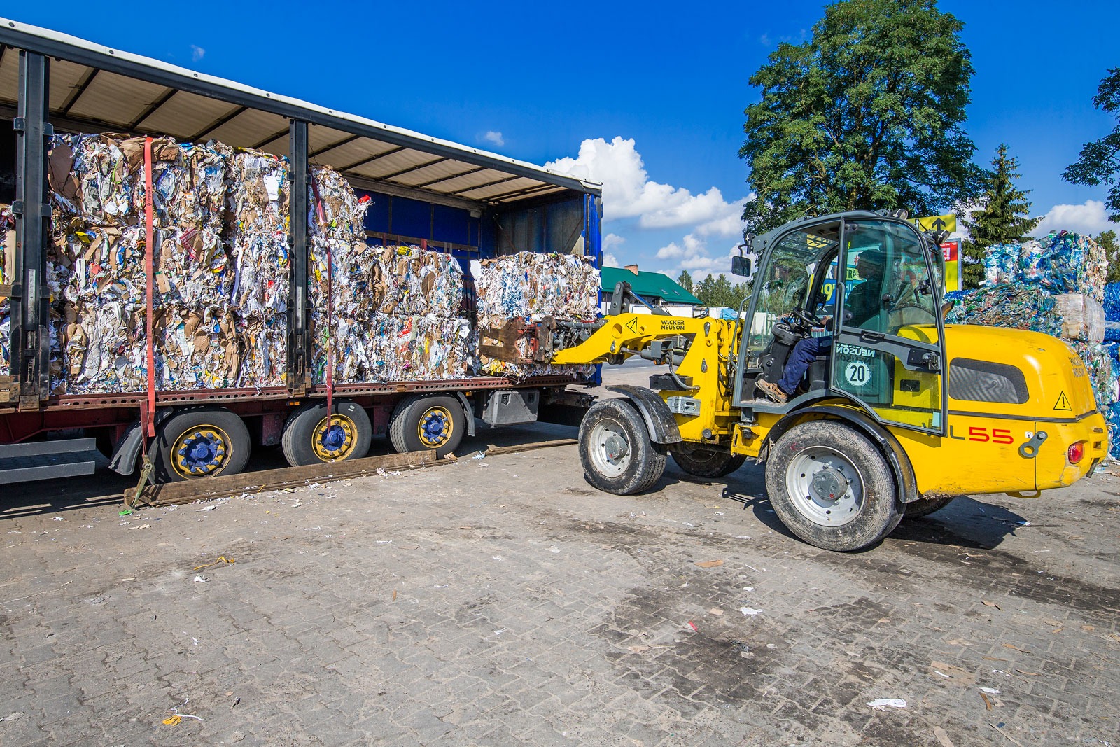 Żółta ładowarka teleskopowa Wacker Neuson L55 podnosi sprasowane bele makulatury na naczepę ciężarówki na tle błękitnego nieba.