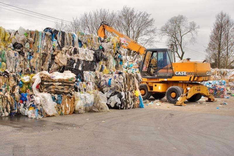 Żółta ładowarka CASE 988 podnosi belę sprasowanych odpadów foliowych na składowisku, widoczne drzewa w tle.