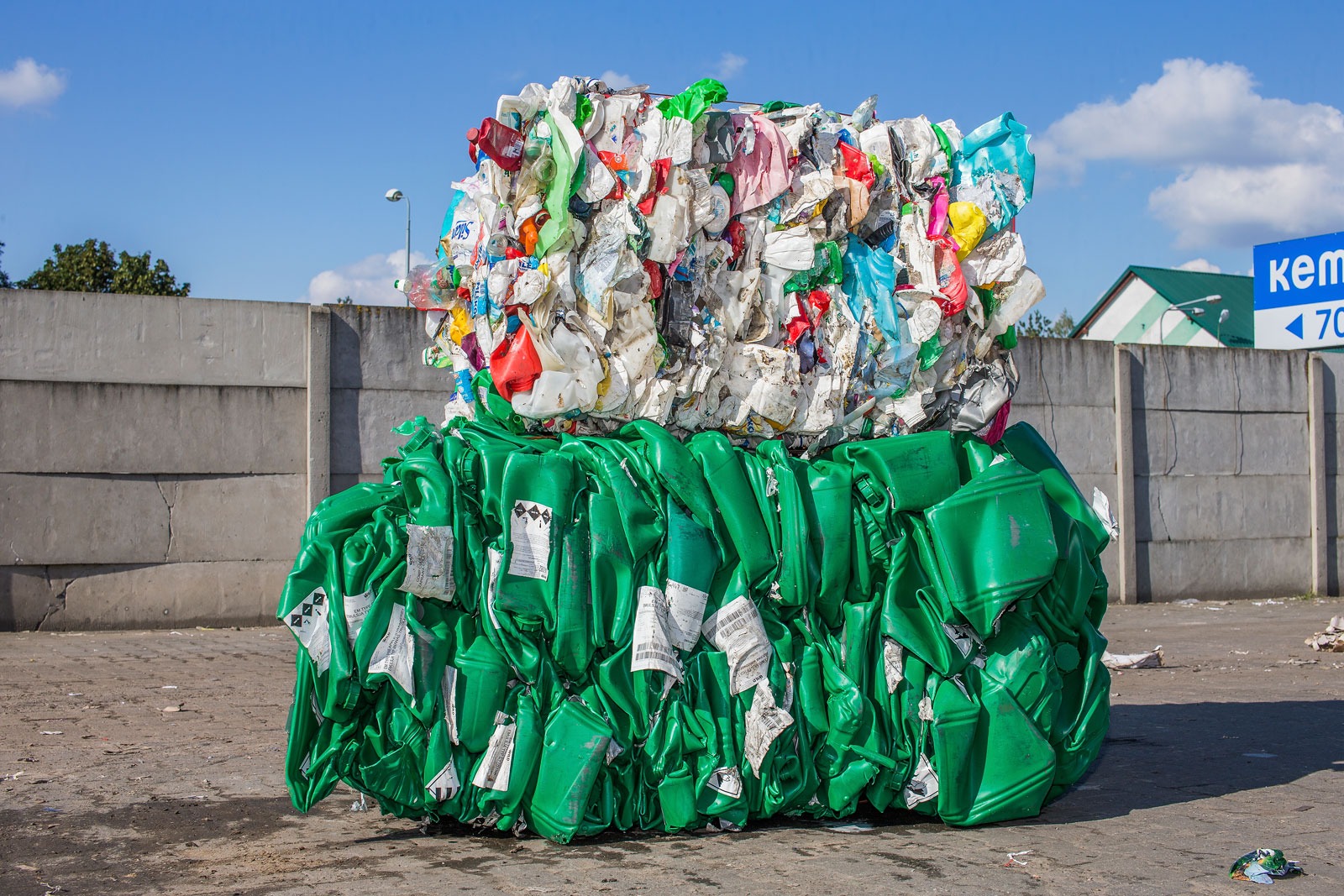 Dwie sprasowane bele odpadów: dolna z zielonych plastikowych kanistrów z etykietami ostrzegawczymi, górna z różnokolorowych, zgniecionych plastikowych opakowań, na tle betonowego muru i błękitnego...