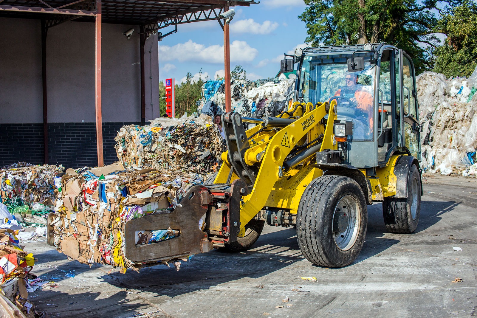 Żółta ładowarka Wacker Neuson transportuje sprasowane odpady kartonowe na tle składowiska śmieci i budynku z zadaszeniem.