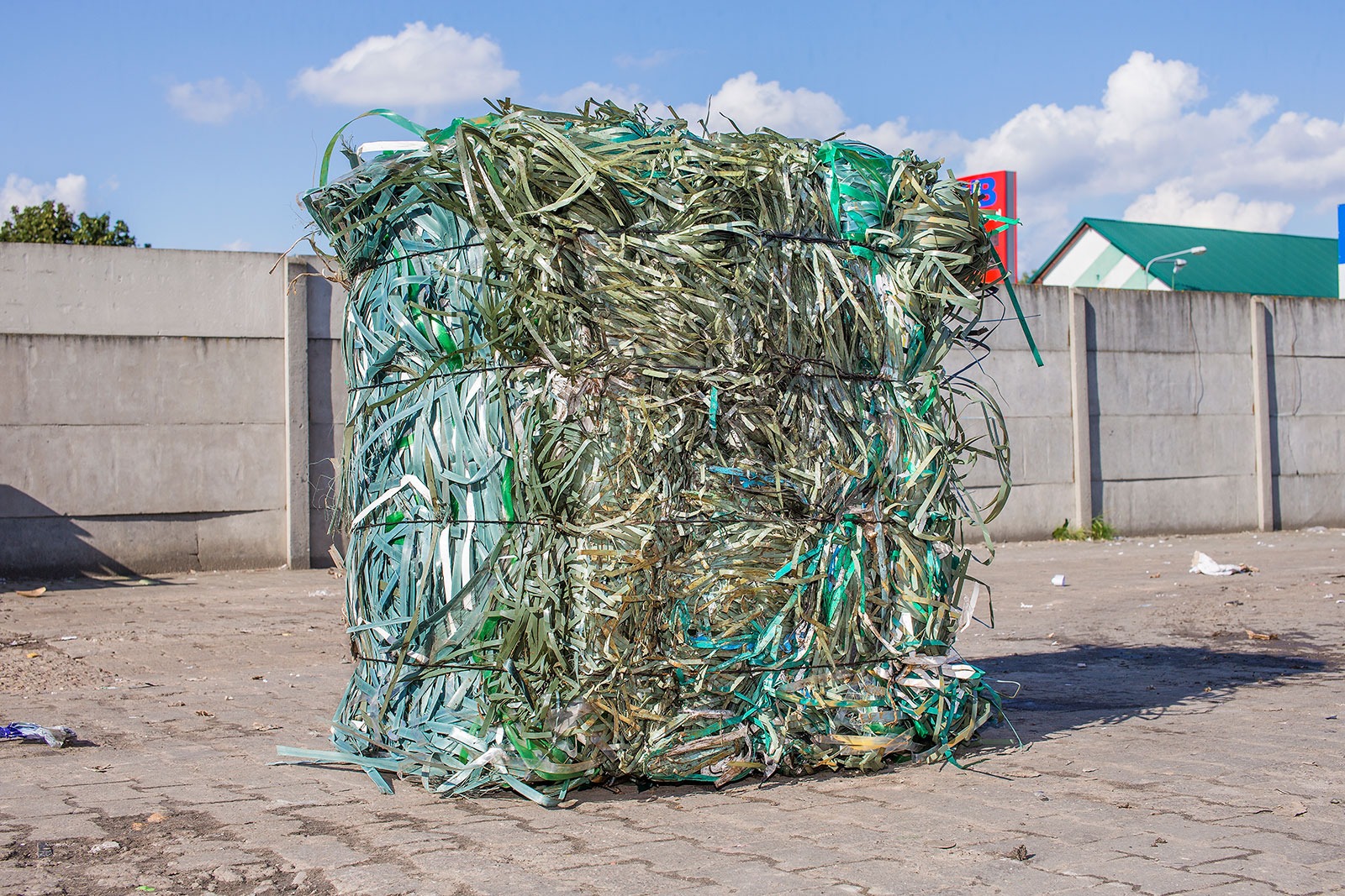 Zbita w kostkę, sprasowana folia budowlana w odcieniach zieleni i bieli, zabezpieczona czarnym sznurkiem, na tle betonowego muru i fragmentu budynku z zielonym dachem.