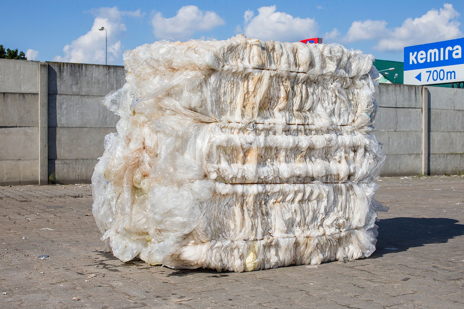 Uformowana w kostkę, sprasowana folia, prawdopodobnie po budowie, składowana na betonowym placu, widoczny betonowy mur i znak drogowy w tle.