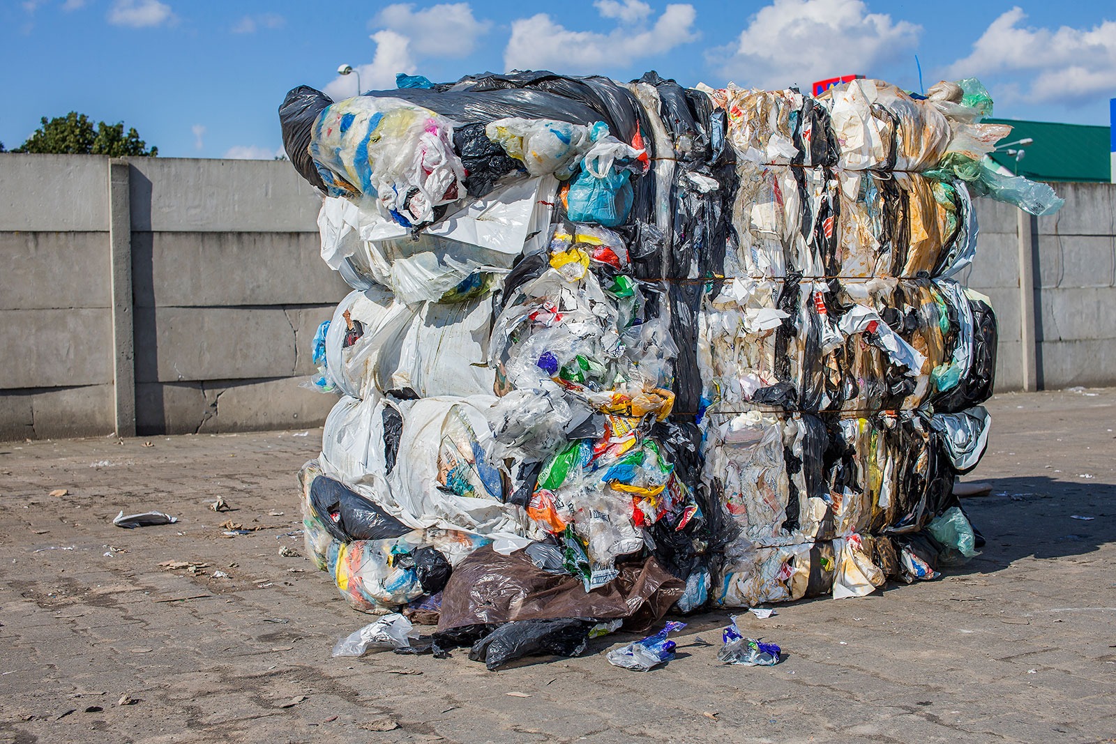 Uformowany w kostkę, sprasowany stos odpadów foliowych i plastikowych, zabezpieczony sznurami, leżący na betonowym placu, z betonowym murem i błękitnym niebem w tle.