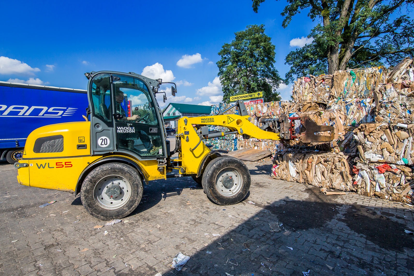 Żółta ładowarka Wacker Neuson WL55 podnosi belę sprasowanej makulatury na placu składowym, w tle niebieska naczepa TIR.