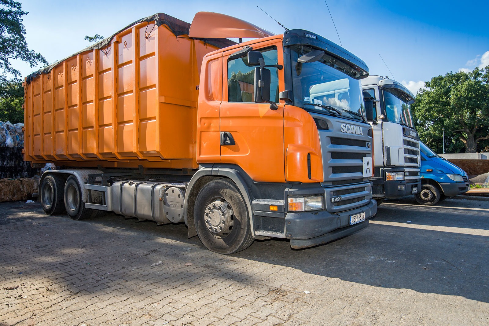 Pomarańczowa ciężarówka Scania z kontenerem, zaparkowana na brukowanej nawierzchni obok dwóch innych pojazdów, pod błękitnym niebem z lekkimi chmurami.