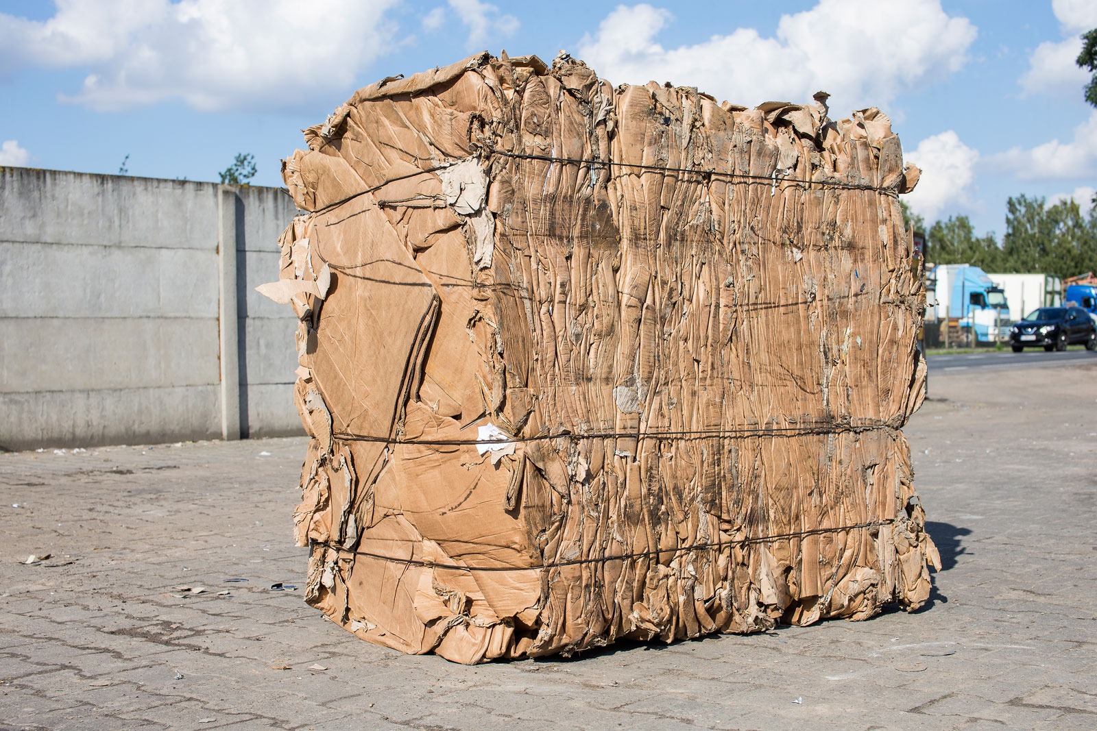 Zbita kostka ze sprasowanej tektury, zabezpieczona czarnym sznurkiem, na betonowym placu, w tle betonowy mur i zaparkowane samochody.
