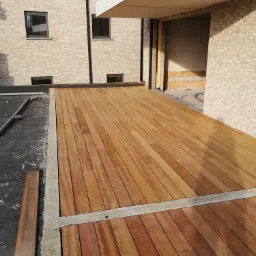 Nowoczesny taras z desek z egzotycznego drewna, z widocznymi śrubami mocującymi, otoczony betonową ramą, w tle budynek w trakcie budowy z cegły.