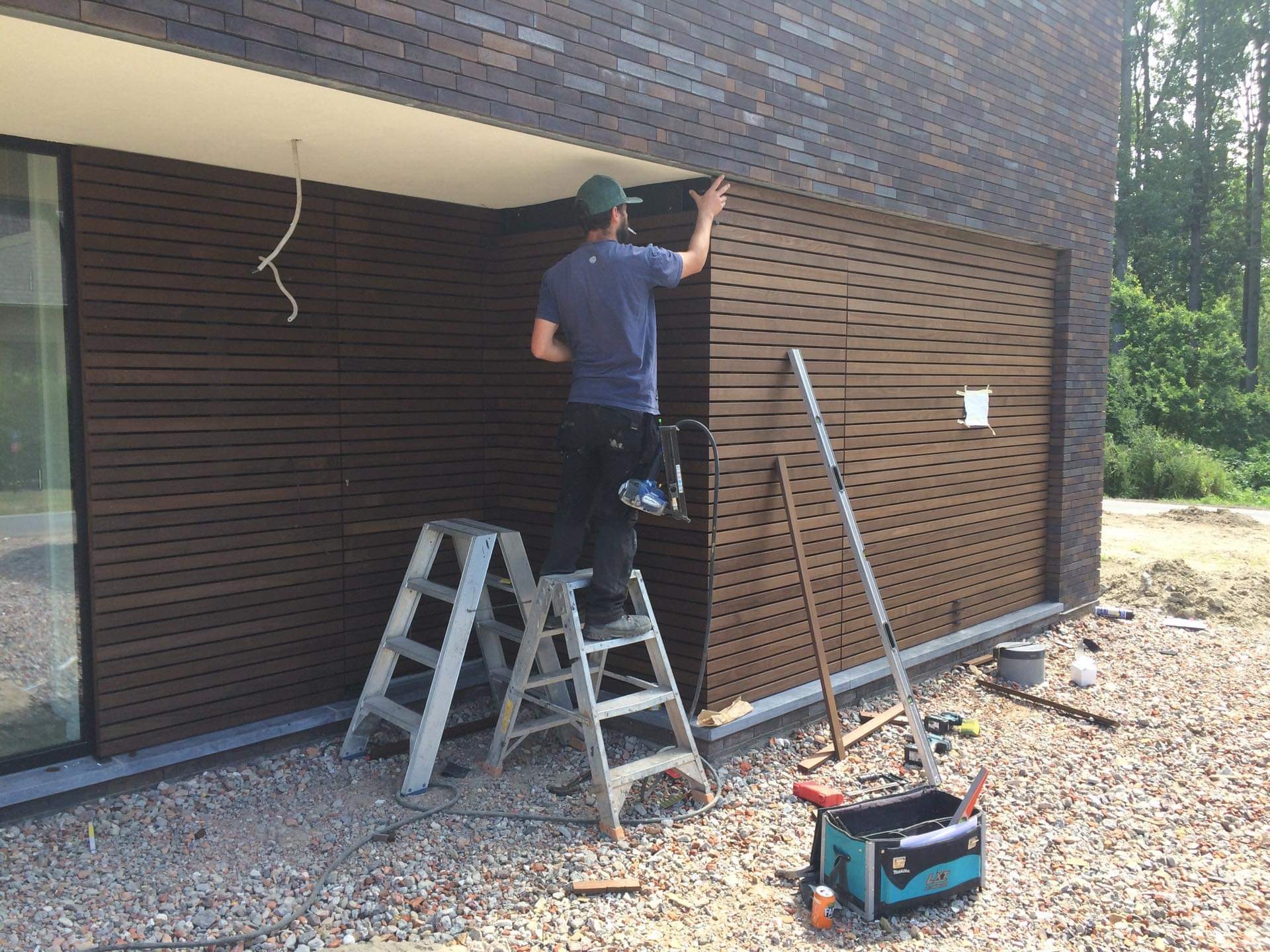 Montaż poziomej, brązowej okładziny elewacyjnej z drewna na ścianie budynku; pracownik na drabinie dopasowuje element, widoczne narzędzia i materiały budowlane na żwirowym podłożu.