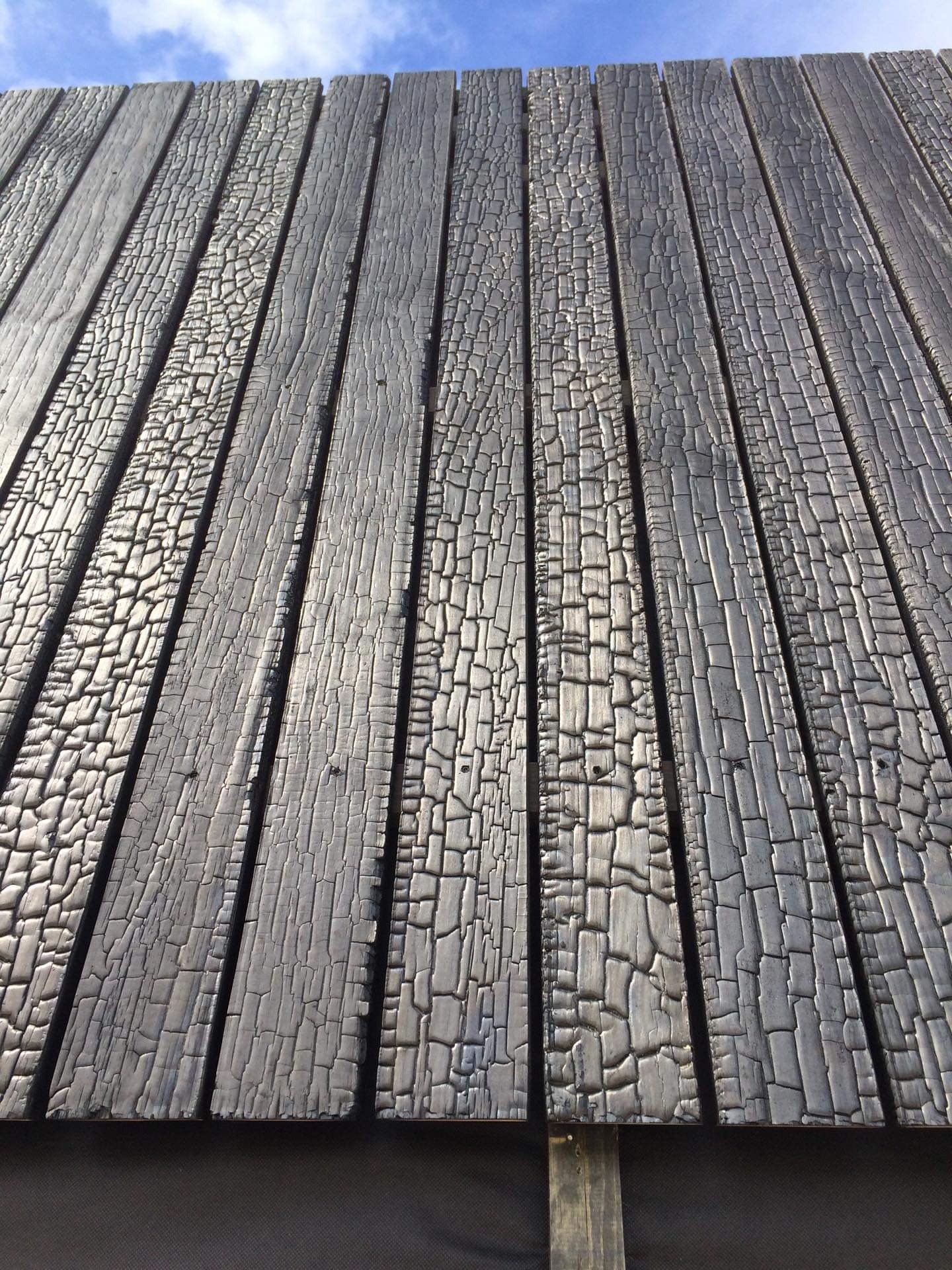 Elewacja z desek opalanych ogniem, tworzących unikalną teksturę spękań i błyszczącej powierzchni, widoczna z dołu pod kątem, na tle błękitnego nieba.