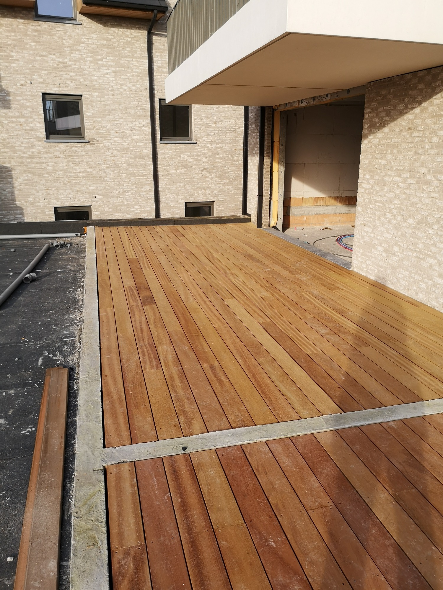 Nowoczesny taras z desek z egzotycznego drewna, z widocznymi śrubami mocującymi, otoczony betonową ramą, w tle budynek w trakcie budowy z cegły.