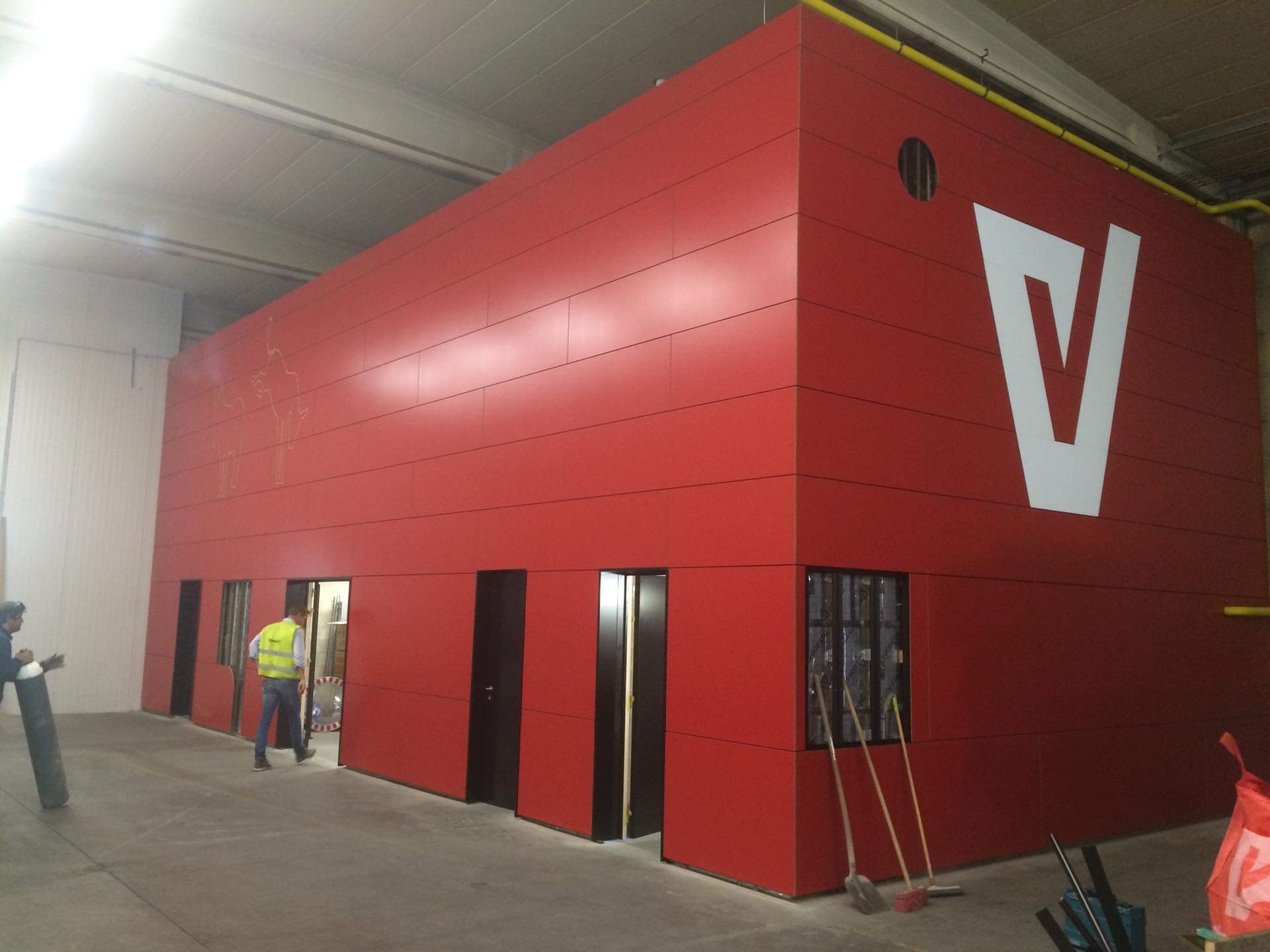 Czerwona konstrukcja z paneli elewacyjnych z białym logo 'V', widoczne drzwi wejściowe i okno z kratami, pracownik w kamizelce odblaskowej wchodzący do środka, w tle narzędzia i butla z gazem.