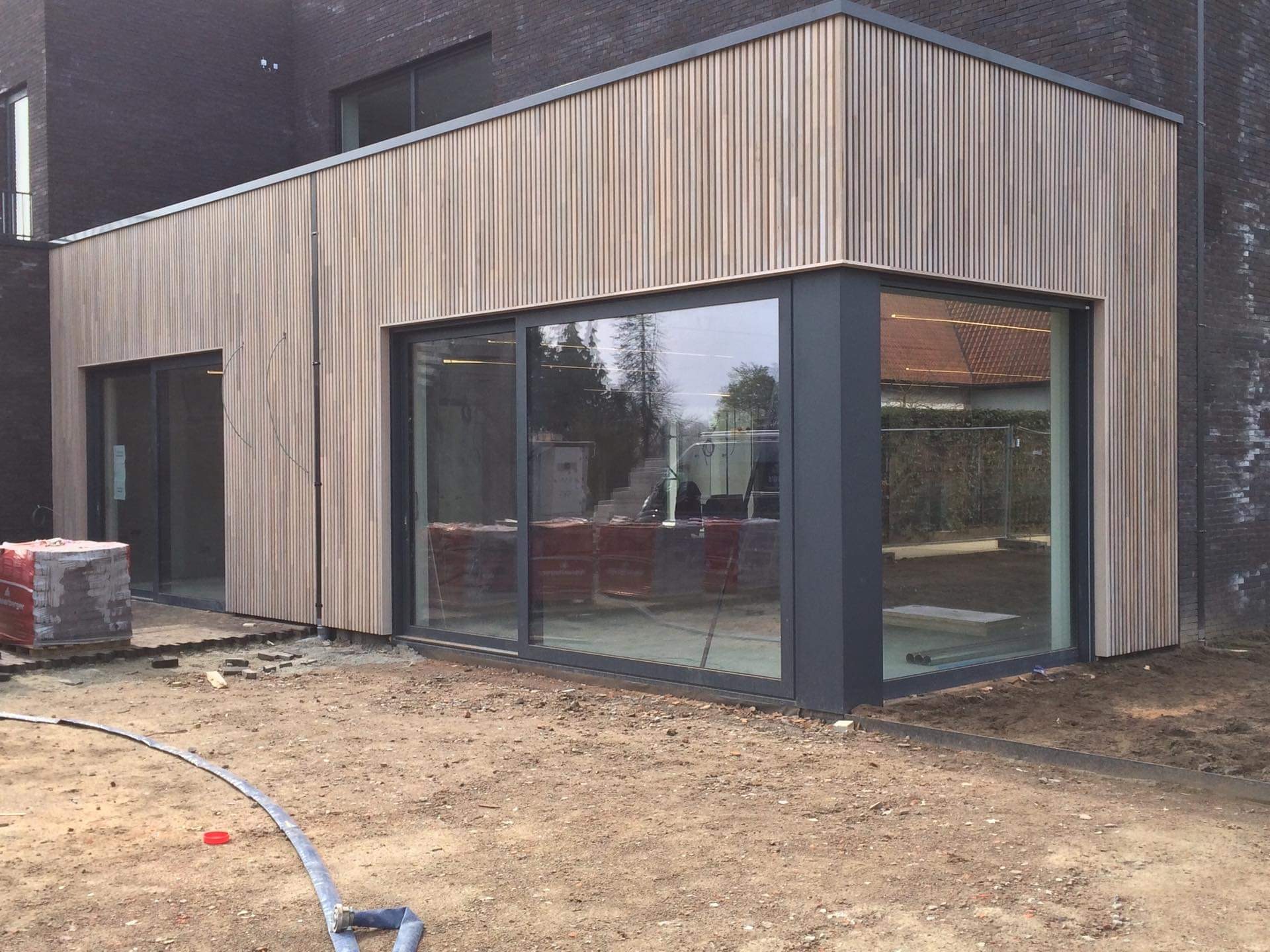 Nowoczesny budynek z elewacją z jasnego drewna w pionowe panele, duże przesuwne okna z ciemnymi ramami, widoczne materiały budowlane i teren wokół w trakcie prac wykończeniowych.