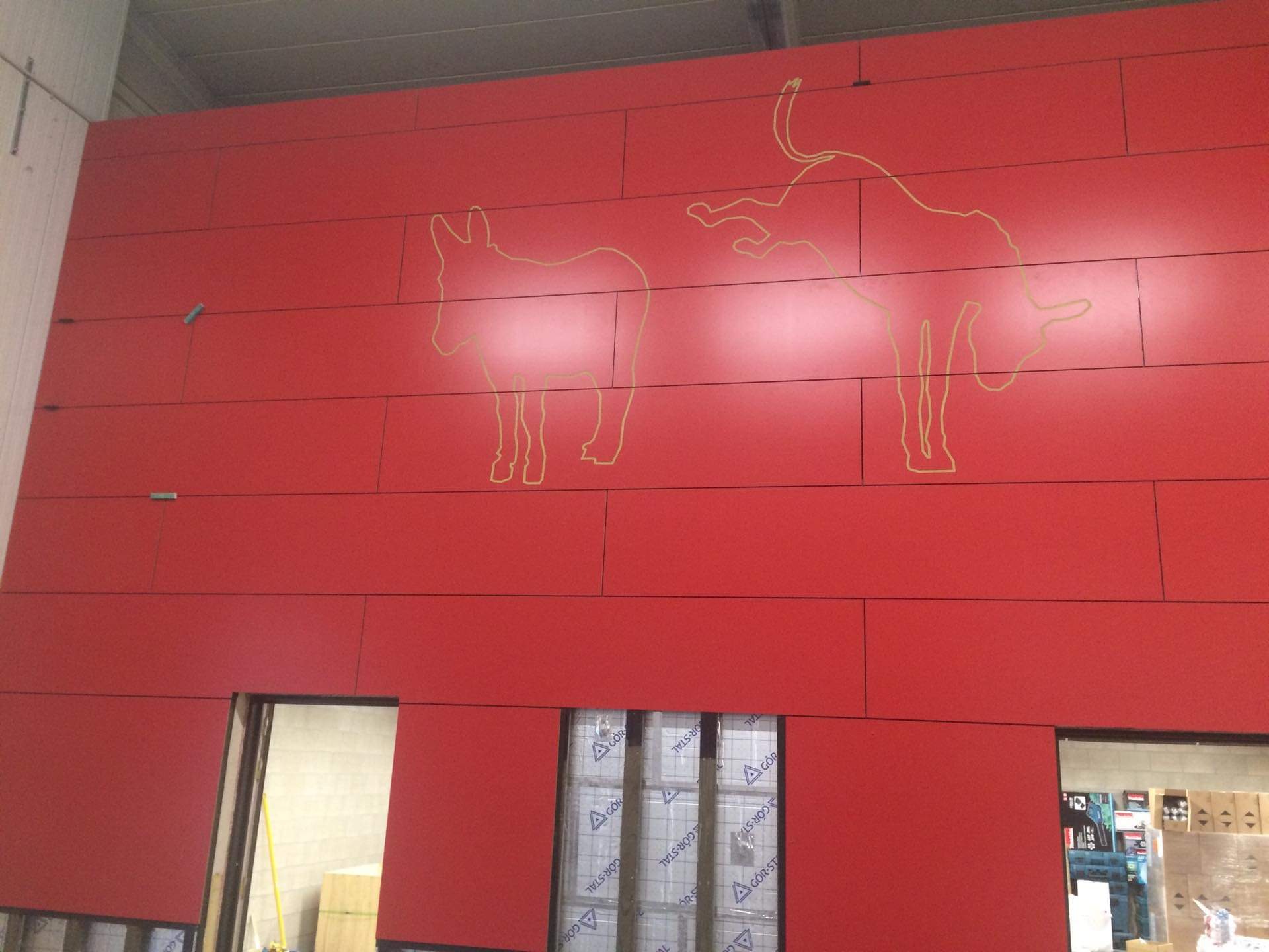 Czerwona elewacja budynku z poziomymi panelami i konturami osła i byka w kolorze żółtym, widoczne otwory okienne i drzwiowe z zabezpieczeniami.