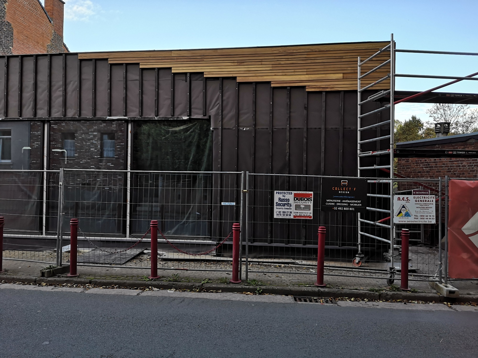 Budynek w trakcie prac elewacyjnych z częściowo zamontowaną drewnianą okładziną, widoczne rusztowania i ogrodzenie budowy.