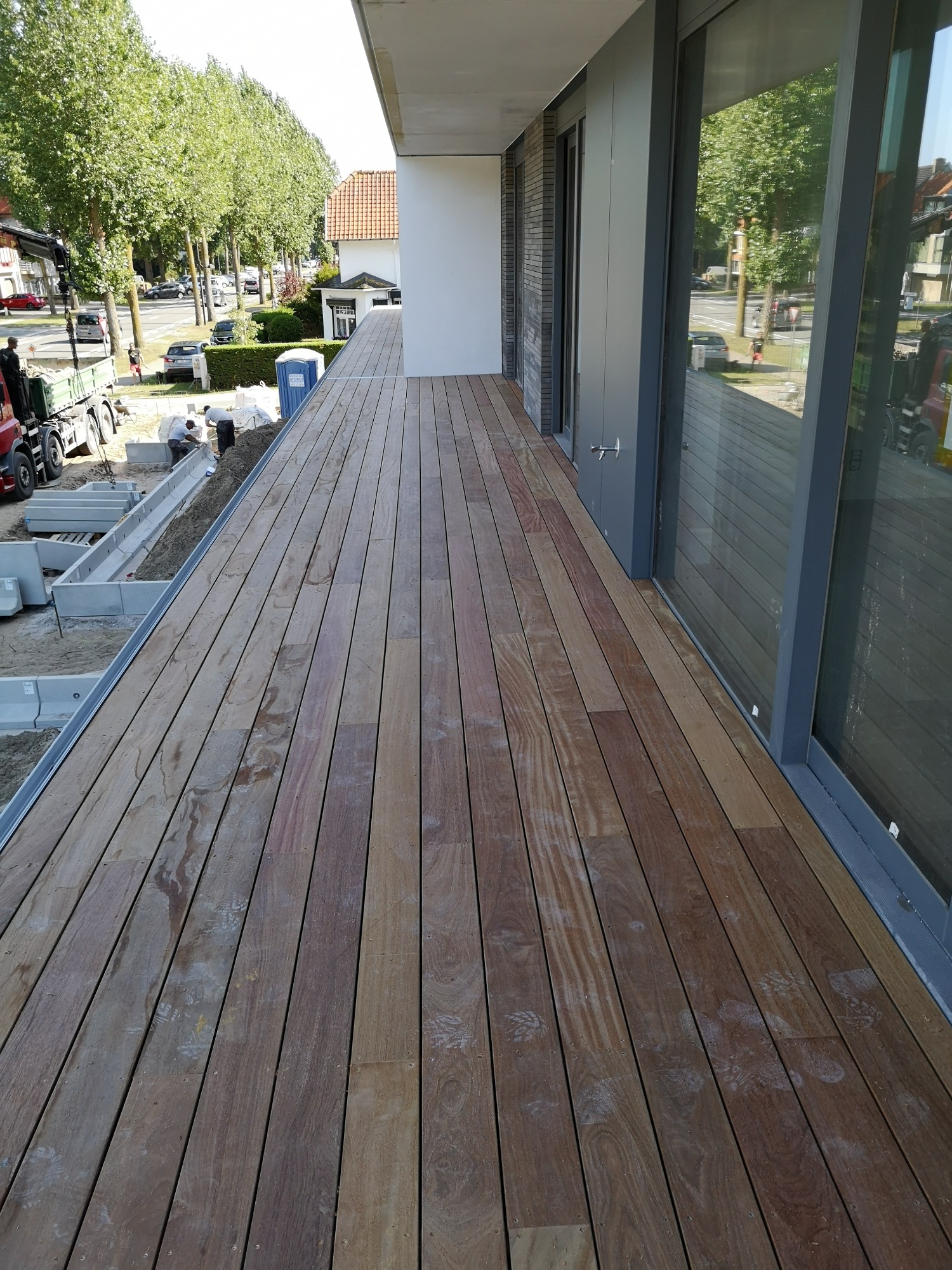 Drewniany taras z widocznymi śladami stóp i zabrudzeniami, przylegający do nowoczesnego budynku z dużymi oknami, w tle prace budowlane i zieleń.