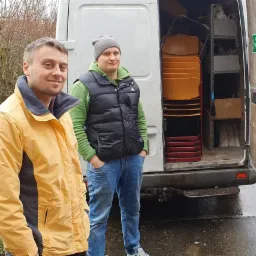 Dwaj mężczyźni stojący obok otwartego białego vana z stosem krzeseł w środku, jeden w żółtej kurtce, drugi w czarnej kamizelce i szarej czapce.