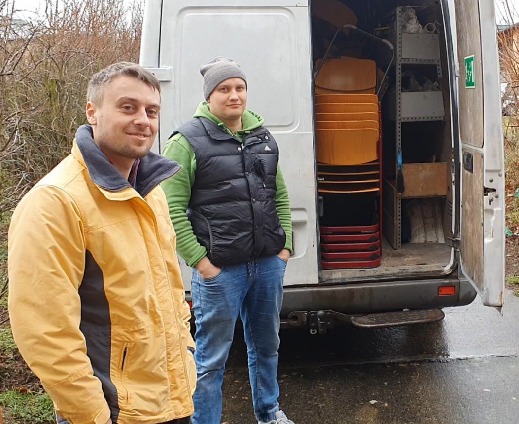 Dwaj mężczyźni stojący obok otwartego białego vana z stosem krzeseł w środku, jeden w żółtej kurtce, drugi w czarnej kamizelce i szarej czapce.
