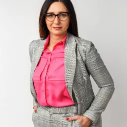 Kobieta w okularach, różowej koszuli i garniturze w kratę pozuje z rękami w kieszeniach na białym tle.