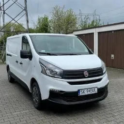 Biały samochód dostawczy Fiat Vivaro zaparkowany na kostce brukowej, widoczna tablica rejestracyjna SK 945UL, w tle garaże i słup wysokiego napięcia.