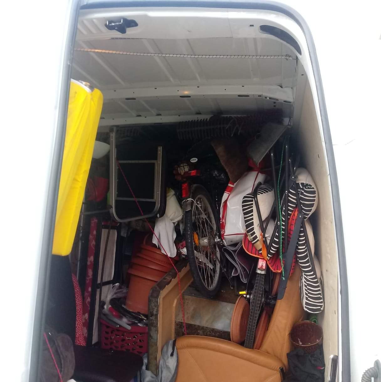 Wnętrze białego busa załadowane różnorodnymi przedmiotami podczas transportu, w tym rower, krzesło, doniczki i worki z rzeczami, widoczne przez otwarte tylne drzwi.