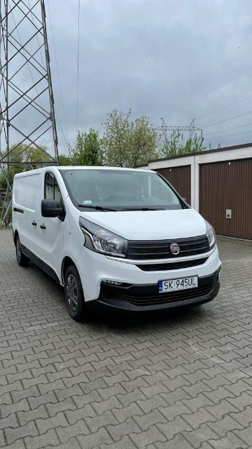 Biały samochód dostawczy Fiat Vivaro zaparkowany na kostce brukowej, widoczna tablica rejestracyjna SK 945UL, w tle garaże i słup wysokiego napięcia.