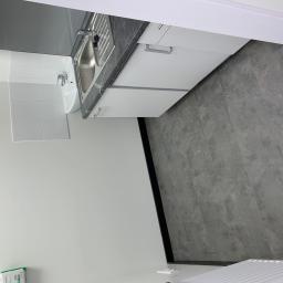 Majessbud Sp. z o.o. - Nowoczesna, minimalistyczna łazienka z białymi szafkami, szarą armaturą i płytkami imitującymi kamień na podłodze, widok na umywalkę i fragment grzejnika.