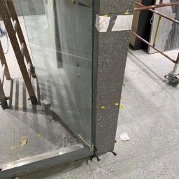 Majessbud Sp. z o.o. - Montaż szklanej ścianki działowej z szarą, kamienną ramą, widoczne podpórki i taśma zabezpieczająca, drabina w tle.