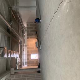 Majessbud Sp. z o.o. - Pracownik w niebieskiej bluzie szpachluje ścianę w surowym wnętrzu, widoczne rury wentylacyjne pod sufitem i ściany przygotowane pod malowanie z widocznymi łączeniami płyt.