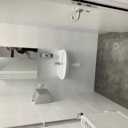 Majessbud Sp. z o.o. - Nowoczesna, biała łazienka z umywalką, lustrem z odbiciem osoby robiącej zdjęcie, grzejnikiem i szarą ścianą wyłożoną płytkami imitującymi beton.