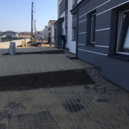 Układanie kostki brukowej na podjeździe przed szeregiem nowych domów, pracownik zamiata piasek, widoczne przygotowane rabaty na trawnik.