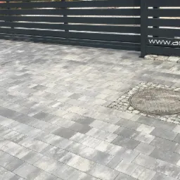 Nowoczesna, przesuwna brama w kolorze antracytowym z poziomymi panelami, widoczna z poziomu brukowanego podjazdu z włazem kanalizacyjnym obłożonym kostką granitową. Na bramie widoczne logo firmy.