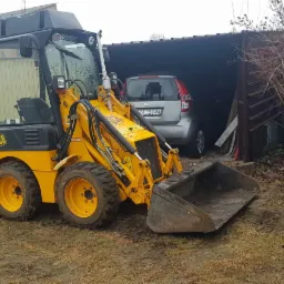 Ż&oacute;łta mini-ładowarka JCB podczas prac ziemnych przy wykopie pod fundament ogrodzenia, widoczni robotnicy i fragment domu w tle.