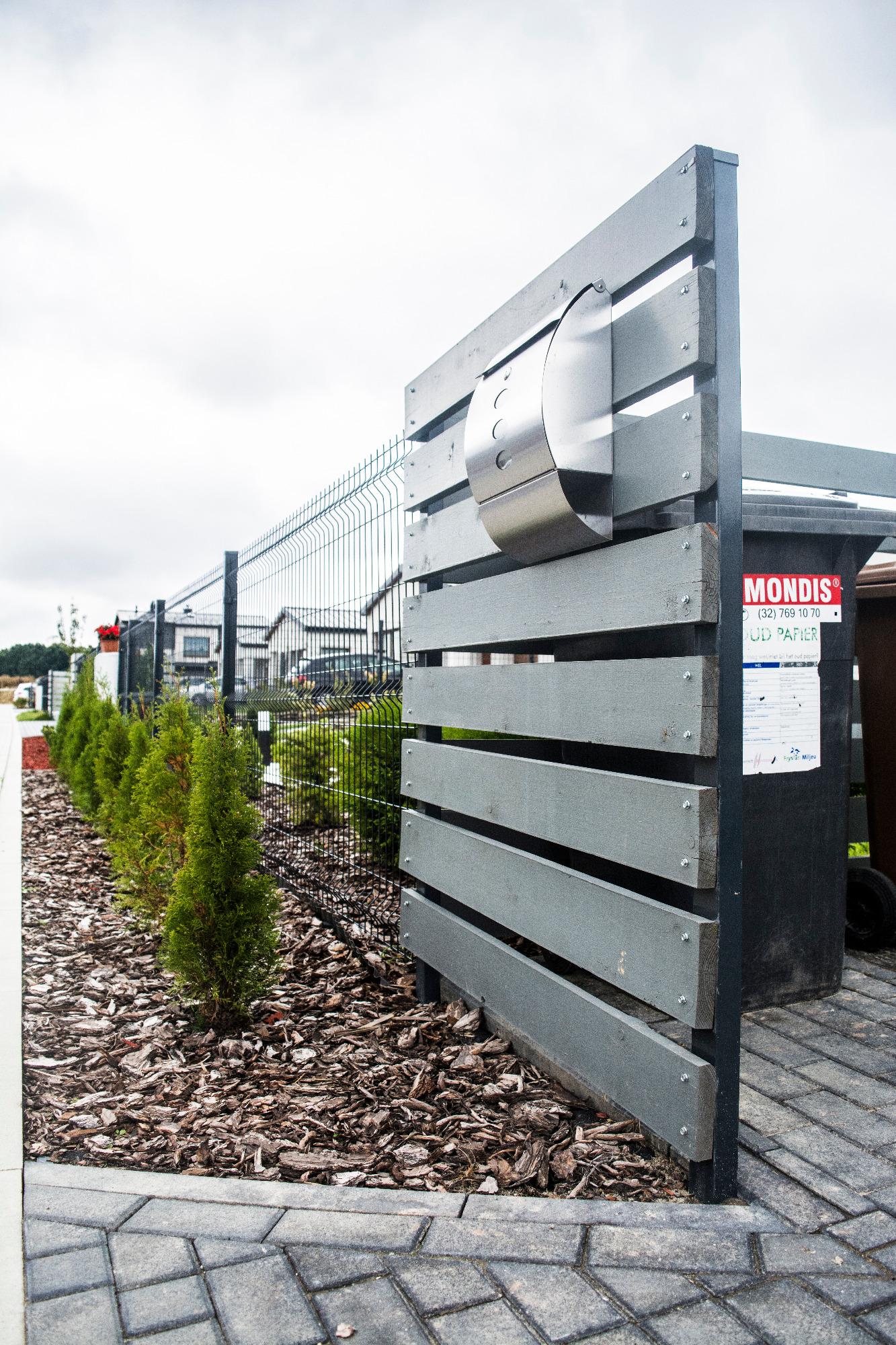 Szara, pozioma, drewniana przesłona z metalową skrzynką na listy, przylegająca do metalowego ogrodzenia panelowego, z młodymi tujami posadzonymi wzdłuż ogrodzenia, na tle domów.