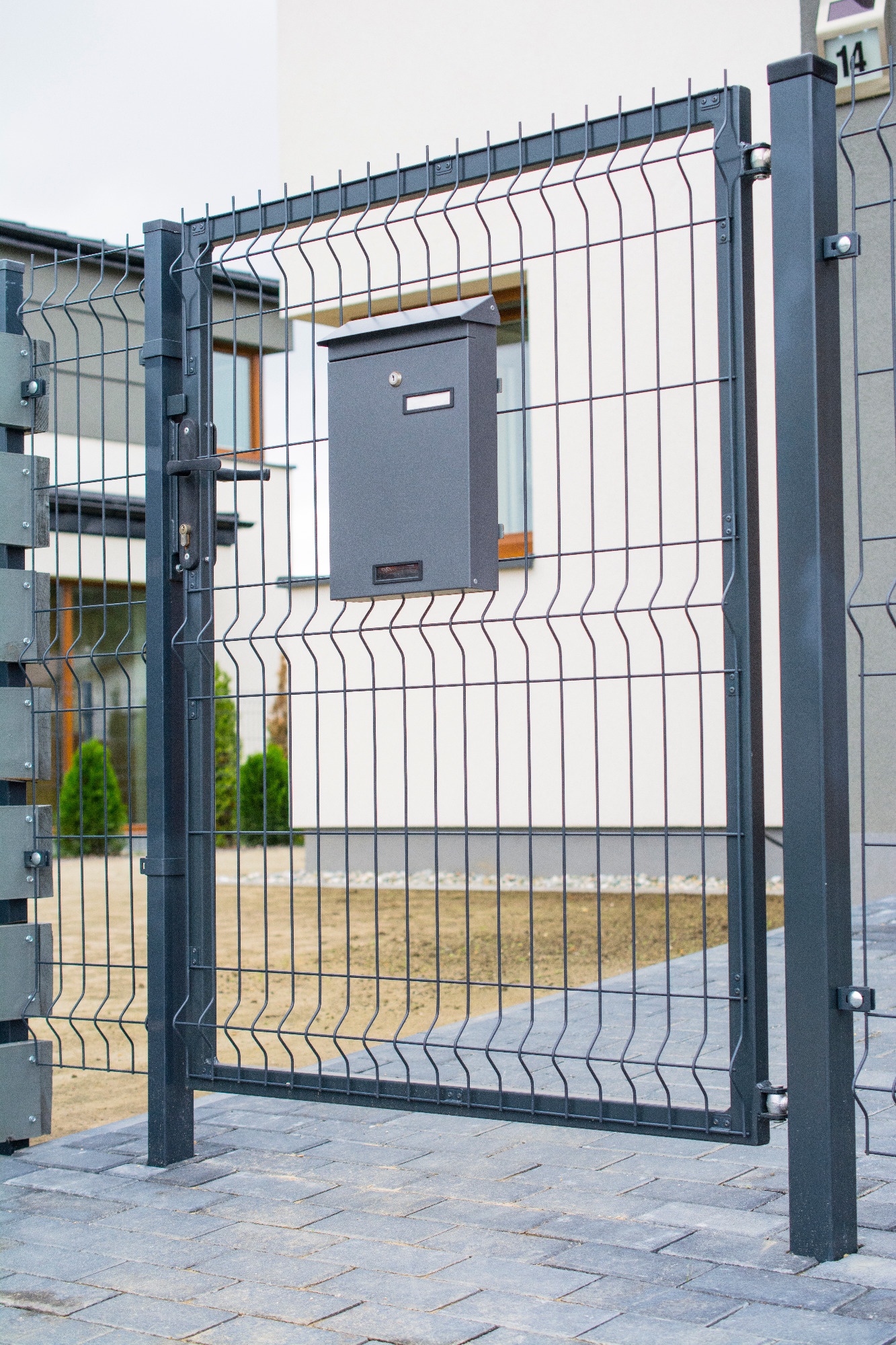 Otwarta, grafitowa furtka z metalowej siatki panelowej, z zamontowaną skrzynką pocztową na tle jasnego budynku.