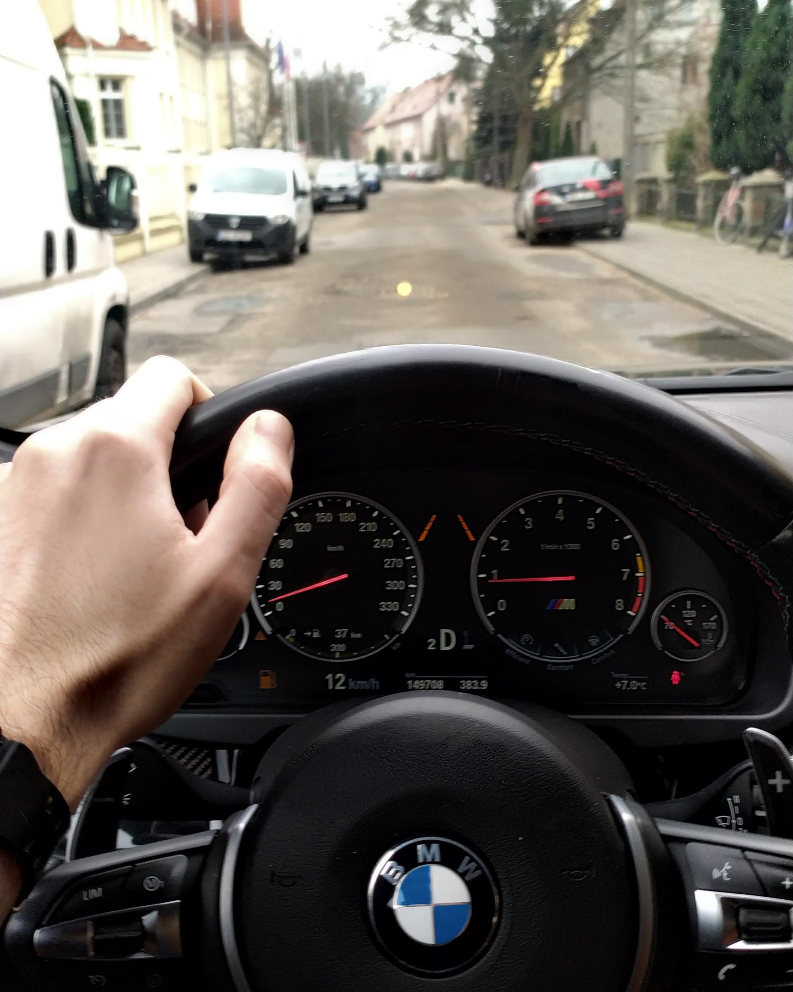 Widok z perspektywy kierowcy BMW na ulicę miejską, skupiający się na desce rozdzielczej, kierownicy z logo BMW i dłoni na kierownicy, z widocznymi budynkami i samochodami w tle.