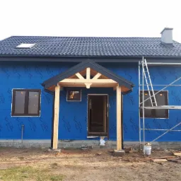 Dom w trakcie budowy z niebieską membraną na ścianach, drewnianym zadaszeniem nad wejściem, czarnym dachem i rusztowaniem z boku.