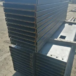 Stos stalowych profili C, przygotowanych do budowy konstrukcji, z widocznymi otworami montażowymi i elementami podkładu, na placu budowy.