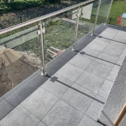 Wykończony taras z szarych płyt w Puławach, z nowoczesną szklaną balustradą ze stali nierdzewnej, widok na ogród z domkiem dla dzieci, obok hałda piasku i taczka.