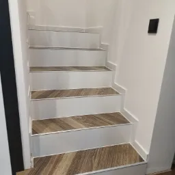 Schody wewnętrzne wykończone panelami imitującymi drewno, z białymi stopnicami i srebrnymi listwami krawędziowymi.