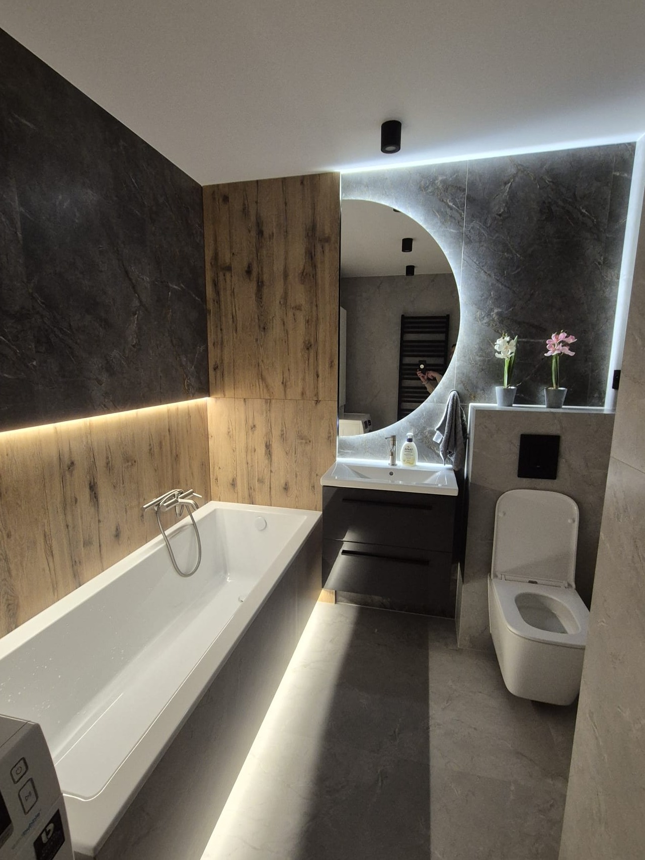 Nowoczesna łazienka z podświetlanym lustrem w kształcie półksiężyca, marmurową ścianą, drewnianymi panelami i białą armaturą, oświetlona taśmą LED pod wanną.