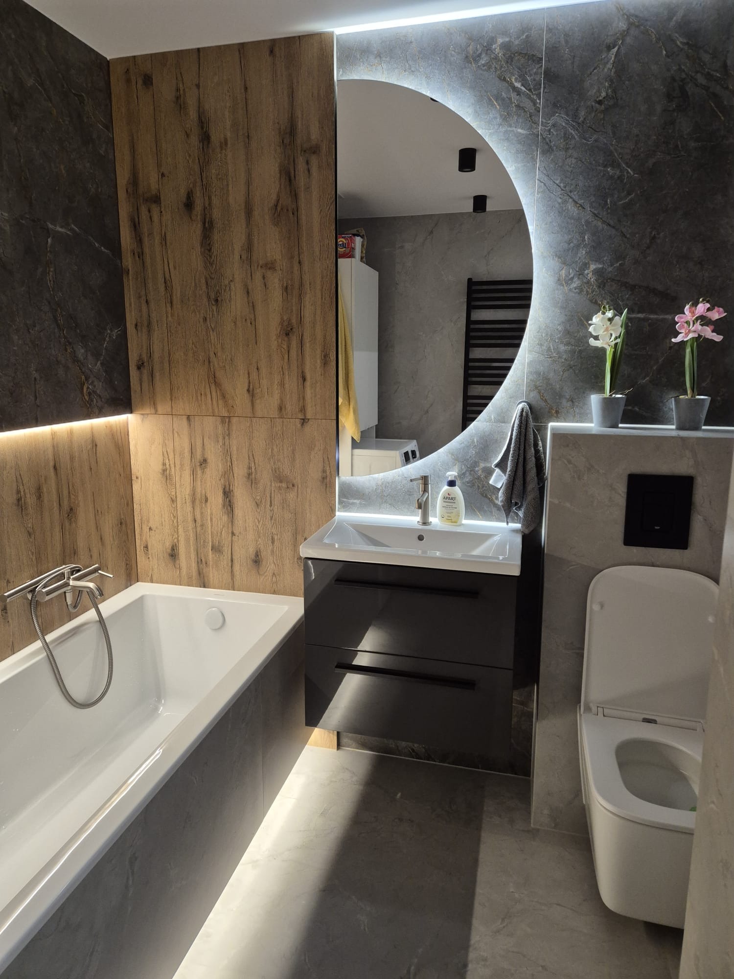 Nowoczesna łazienka z wanną obudowaną szarymi płytkami, umywalką z szafką w kolorze grafitowym, okrągłym lustrem z podświetleniem LED i toaletą; na ścianach połączenie płytek imitujących drewno...