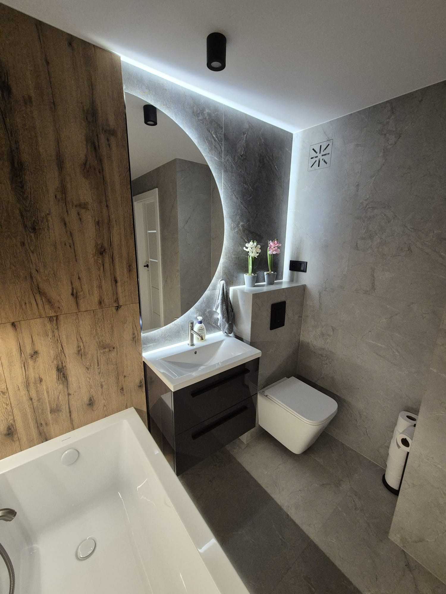 Nowoczesna łazienka z drewnianą okładziną, okrągłym lustrem z podświetleniem LED, umywalką z szafką, podwieszaną toaletą i szarą płytką imitującą kamień.
