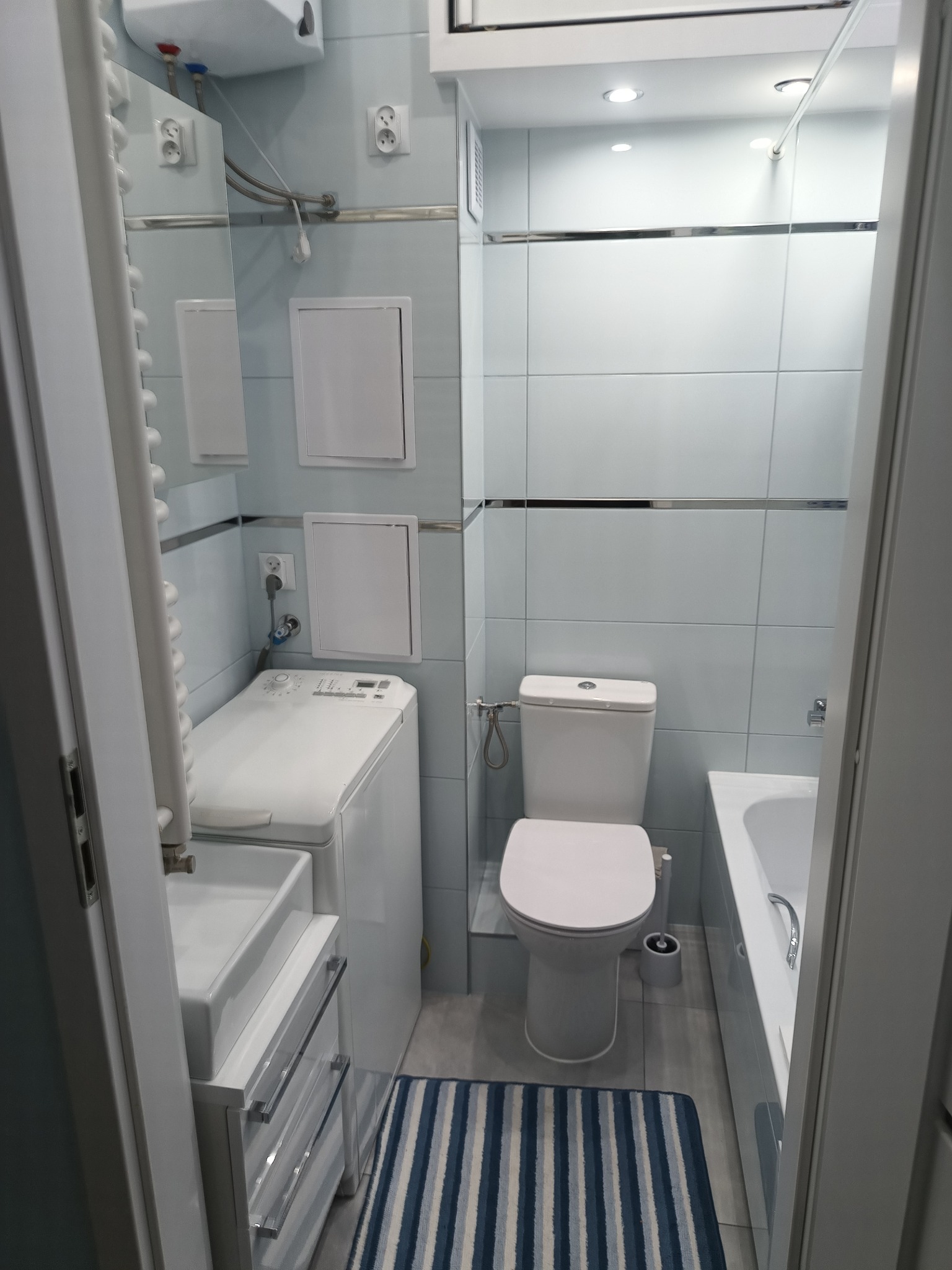 Wnętrze wyremontowanej łazienki z pralką, umywalką z szafką, toaletą i wanną, wykończonej w jasnych płytkach z poziomymi metalowymi listwami dekoracyjnymi.