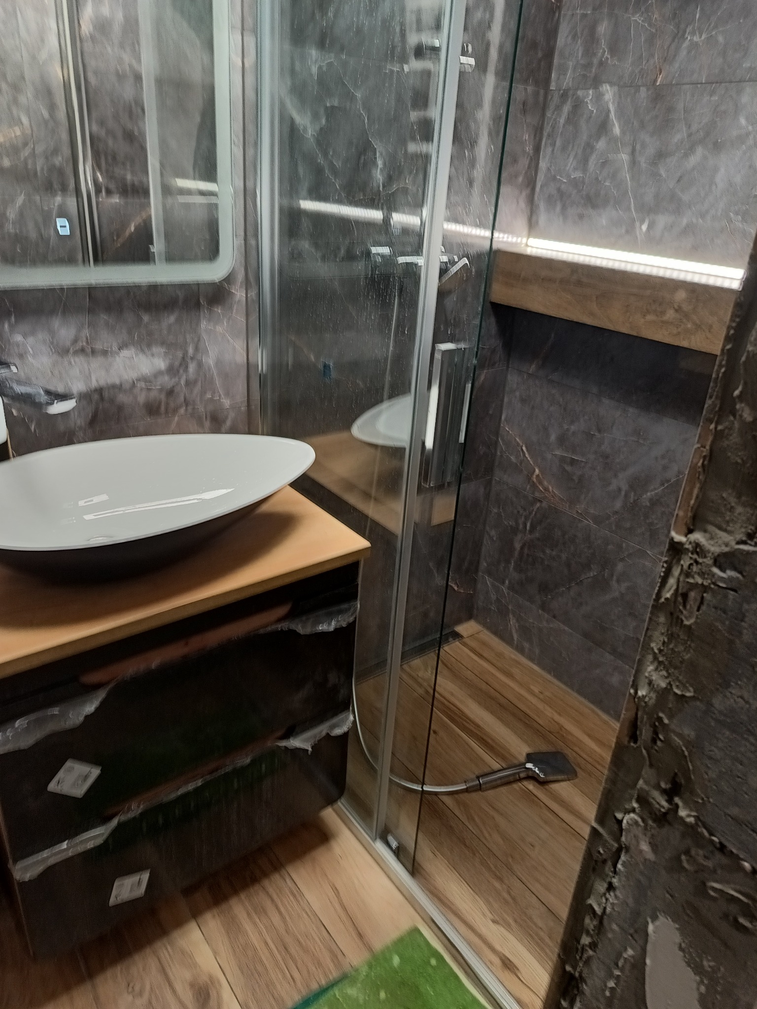 Nowoczesna łazienka z umywalką nablatową, kabiną prysznicową typu walk-in z odpływem liniowym i szarymi płytkami imitującymi marmur, oświetlona listwą LED.