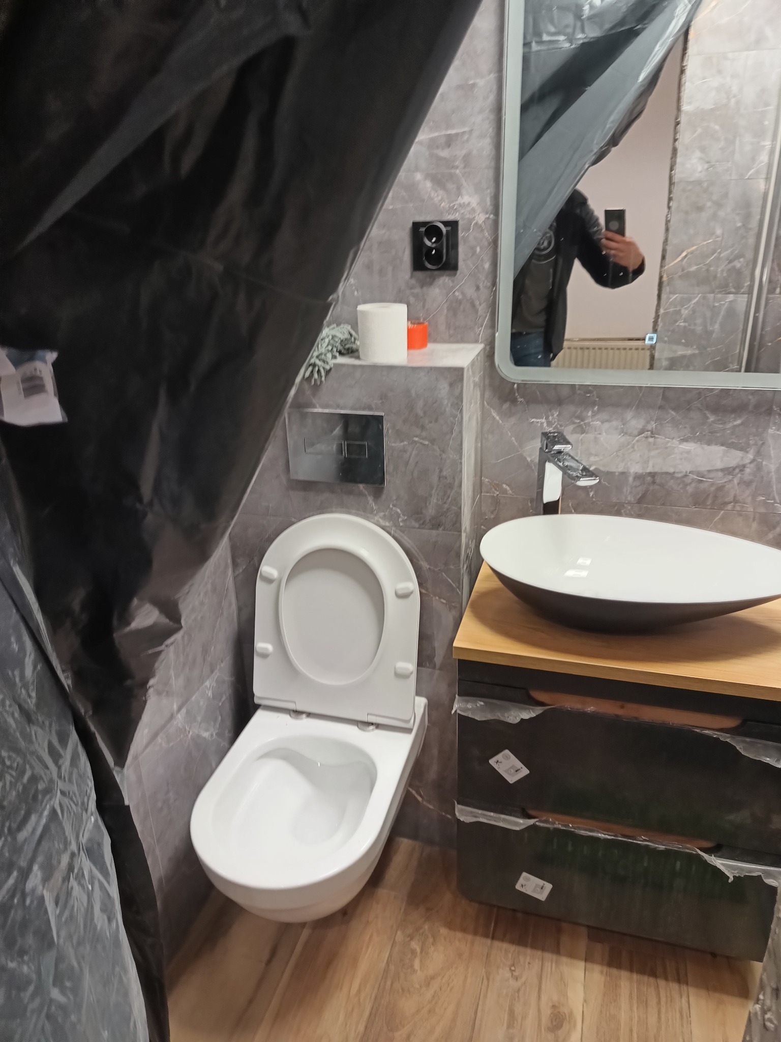 Łazienka w trakcie zabezpieczania przed pracami wykończeniowymi, widoczna toaleta, umywalka nablatowa i lustro, wszystko osłonięte folią malarską.