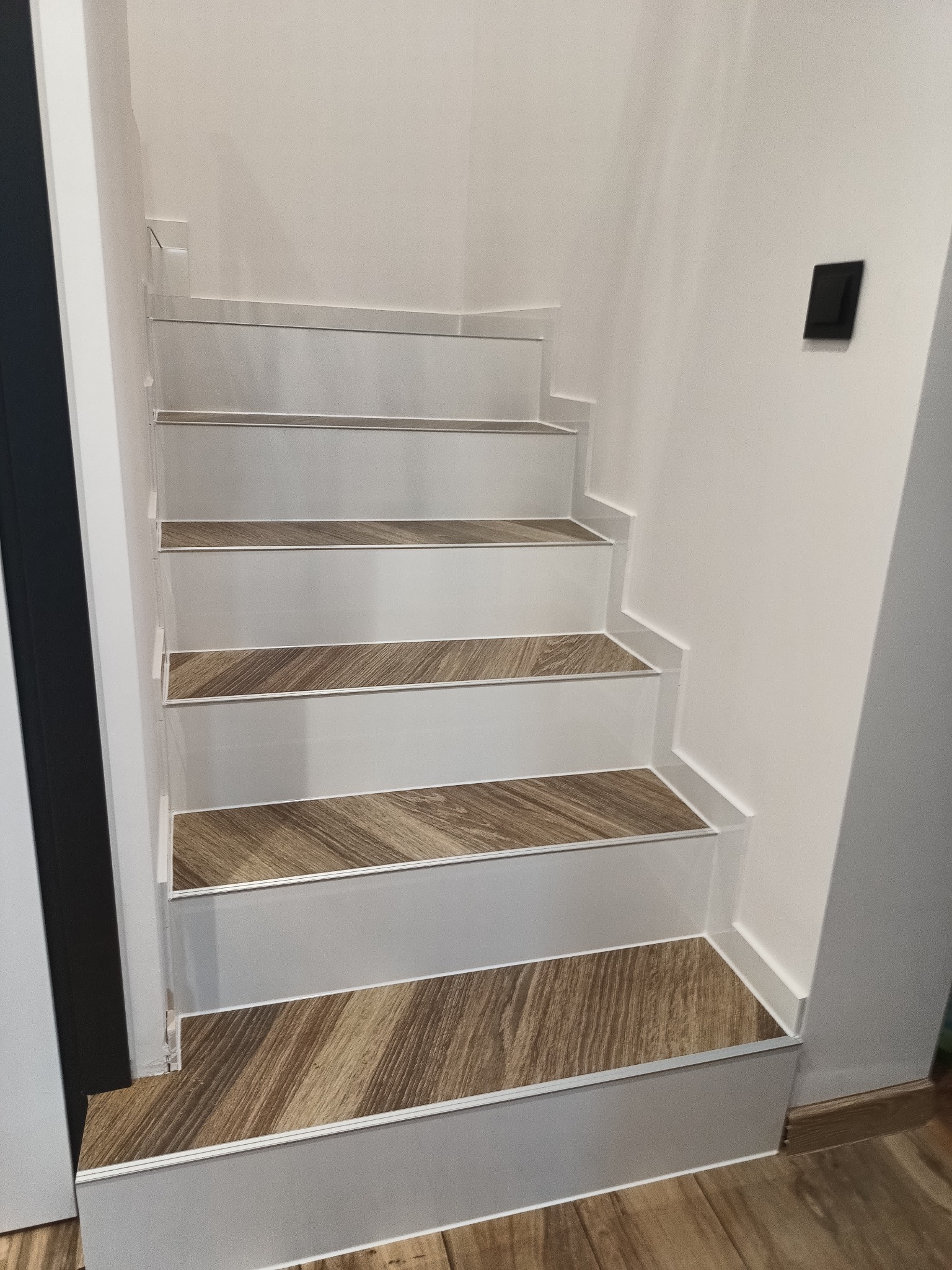 Schody wewnętrzne wykończone panelami imitującymi drewno, z białymi stopnicami i srebrnymi listwami krawędziowymi.