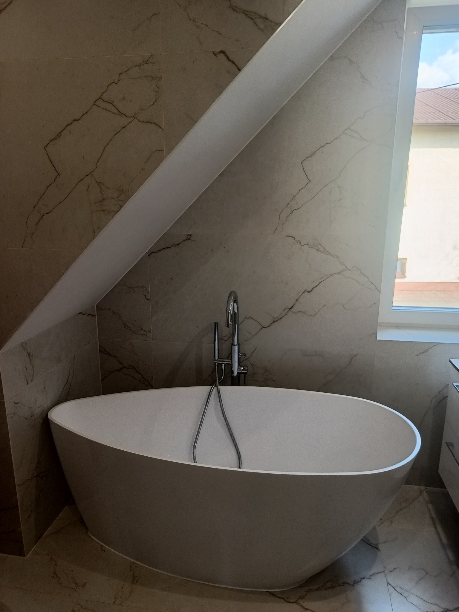 Wolnostojąca wanna w łazience z marmurowymi płytkami, widoczna bateria wannowa z wężem, okno z widokiem na dom z brązowym dachem.