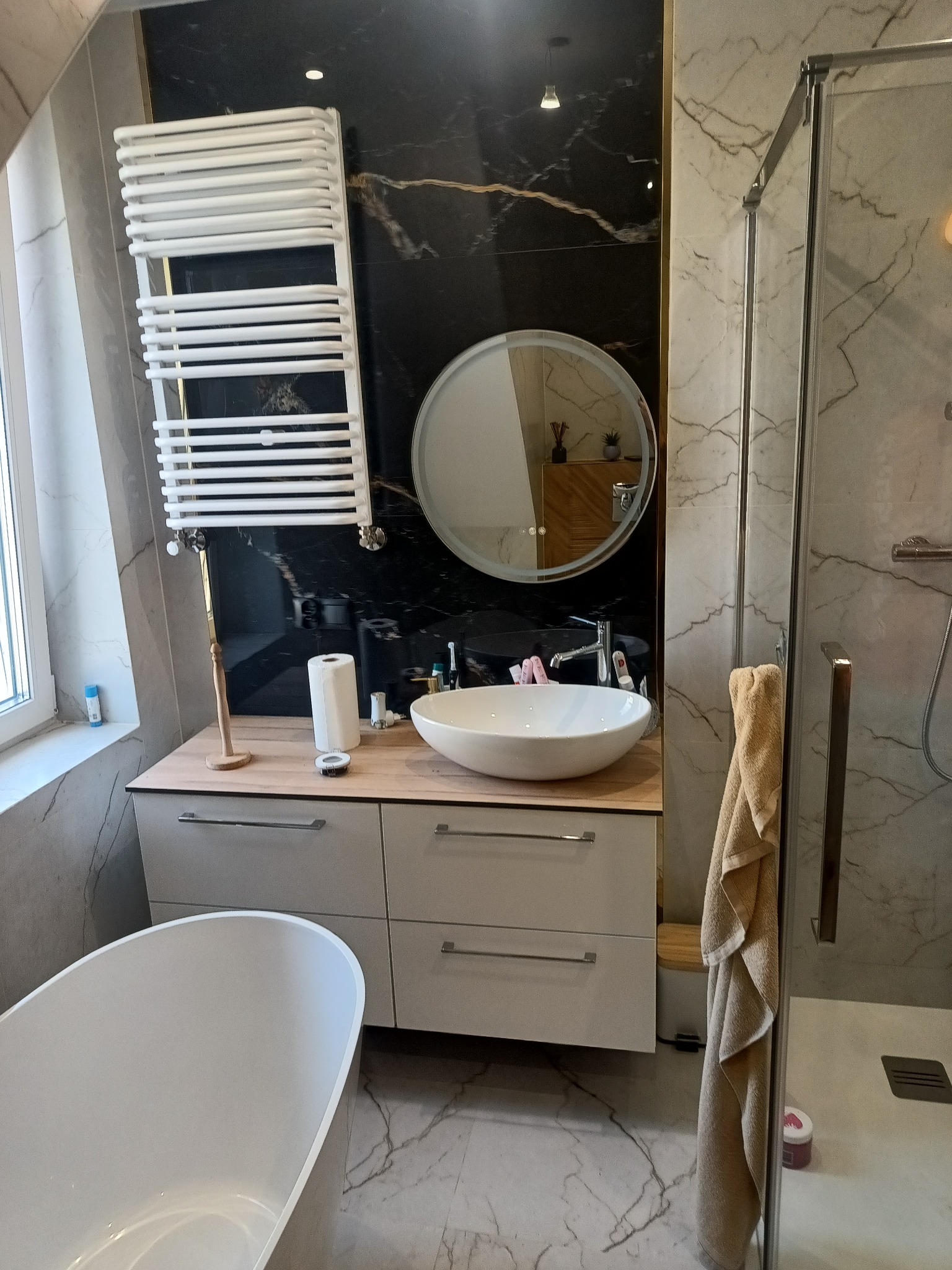 Nowoczesna łazienka z czarno-białymi marmurowymi płytkami, owalną umywalką na szafce z szufladami, okrągłym lustrem z podświetleniem i grzejnikiem drabinkowym.