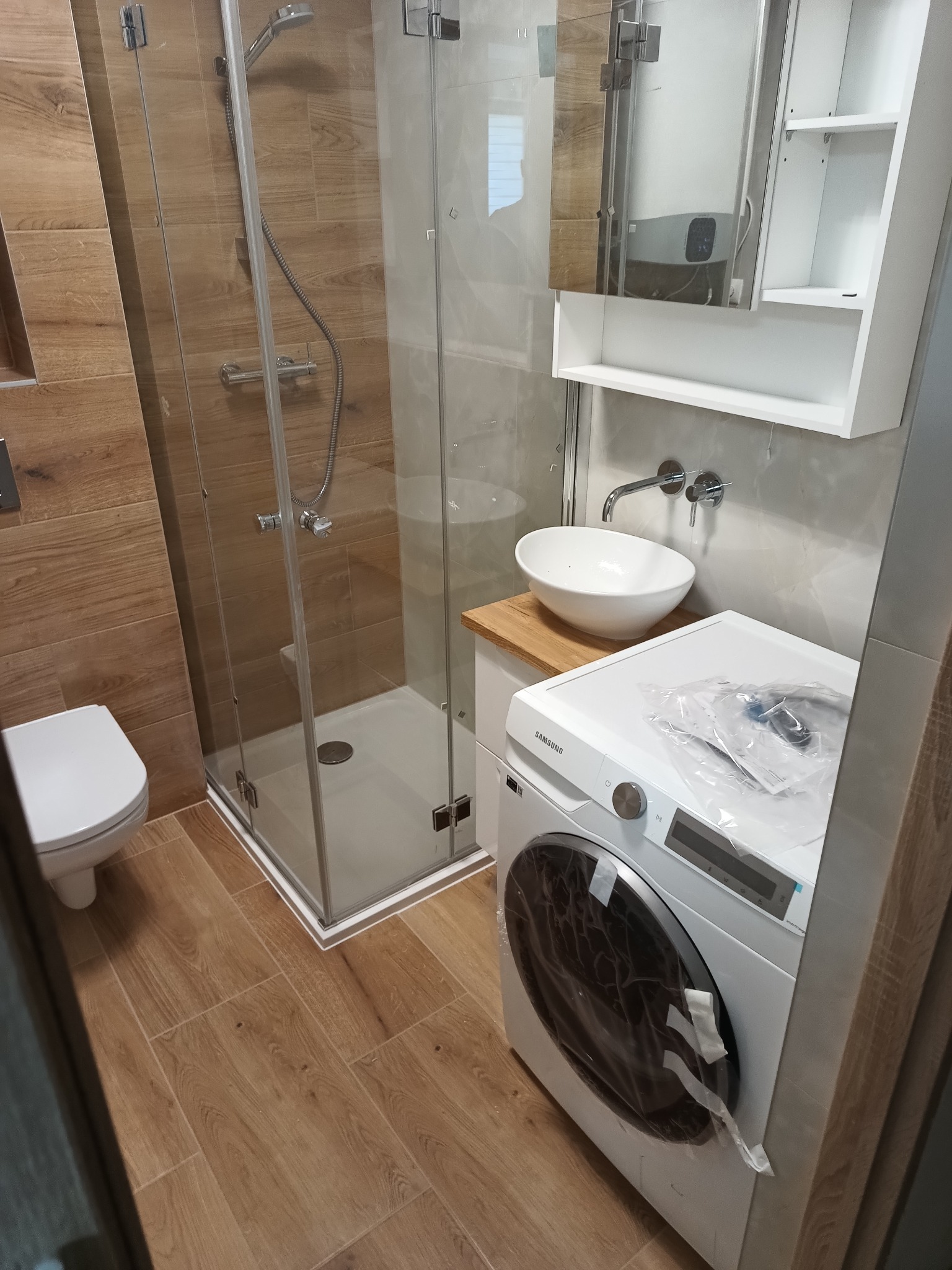 Nowoczesna łazienka z kabiną prysznicową, umywalką nablatową na drewnianym blacie i pralką Samsung jeszcze w folii ochronnej, drewnopodobne płytki na ścianach i podłodze.