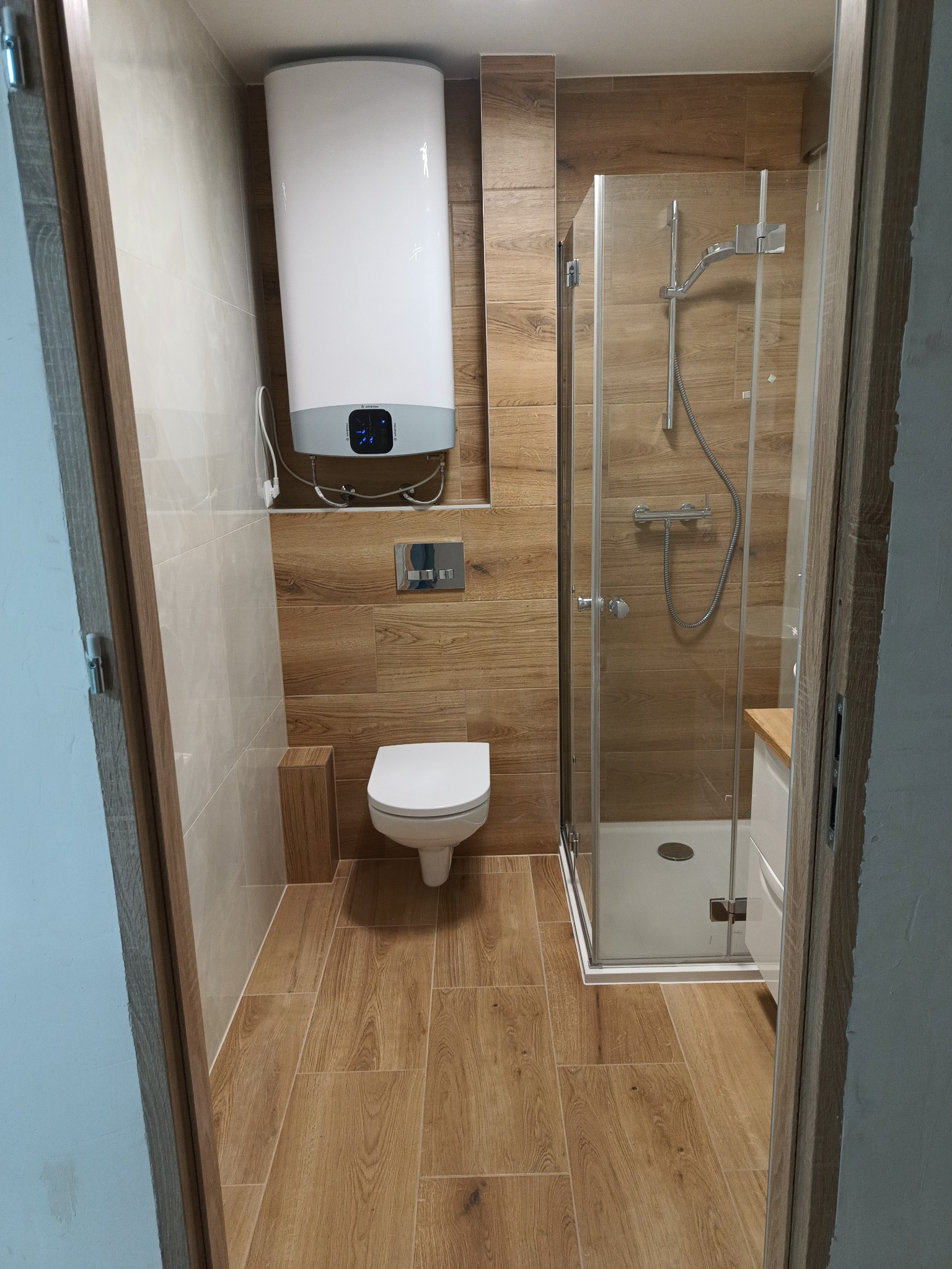 Wykończona łazienka z kabiną prysznicową, toaletą wiszącą i bojlerem Ariston, wykończona płytkami imitującymi drewno na ścianach i podłodze, widok zza uchylonych drzwi.